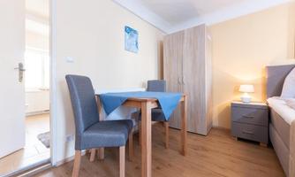 A simple, bright room with a table and two chairs. There is a wardrobe and a cozy bedside lamp.