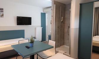A modern hotel room with two single beds, a table, and a television. The decor is simple and bright, with a bathroom area in the corner.