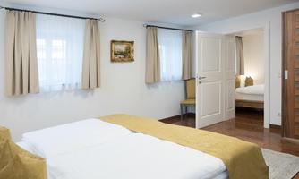 A bright and modern bedroom with two beds and a cozy sitting area. The windows are adorned with white curtains and there is an additional room in the background.