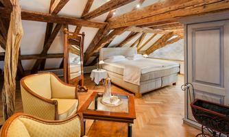 A cozy attic room with wooden beams and a large bed. The furniture consists of a chair, a table, and a mirror.