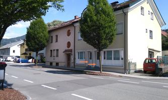A street-side view of a residential building with large windows and two tall trees. Cars are parked on the other side of the street.