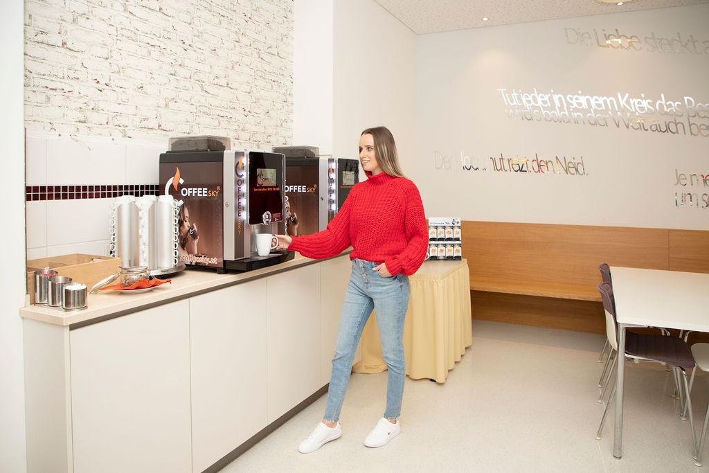 A woman in a red sweater stands by a coffee machine. In the background, more coffee accessories and a cozy seating area can be seen.