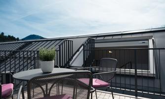 A balcony with a round table and chairs, surrounded by a modern environment. In the background, mountains and a clear sky are visible.