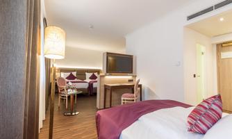 A modern hotel room with a double bed and light wooden floor. The furnishings include a TV, two chairs, and a table.