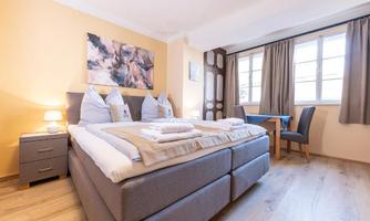 A modern bedroom with a double bed and two pillows. The room has large windows, a table, and a bright, friendly color scheme.