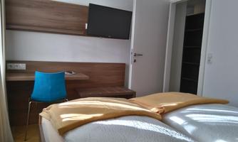 A simple room with a double bed, a desk, and a blue chair. A TV is hanging on the wall.