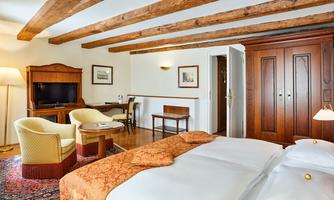 A cozy hotel room with a large double bed and wooden ceilings. There is a television, a desk, and comfortable armchairs.