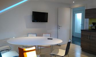 A bright, modern room with an oval table and colorful chairs. A television hangs on the wall, and there is an open door to the kitchen.