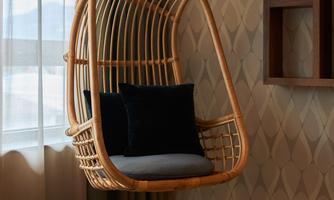 A stylish hanging basket chair with cushions in a cozy room. Behind the chair, a window and wall decor can be seen.