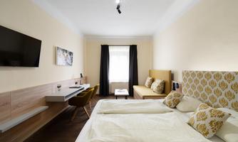 A modern room with a double bed and a cozy seating area. The room is bright with a large window and a television on the wall.