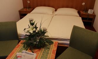 A simple room with a double bed and two chairs. On the table, there is a floral decoration and a newspaper.