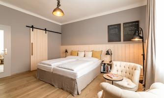 A modern bedroom with two beds, soft colors, and a cozy armchair. The walls are designed simply, and there are decorative wall pictures.
