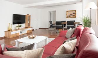A modern living room with red leather furniture and a large television. The room is bright and inviting, with a dining table in the background.
