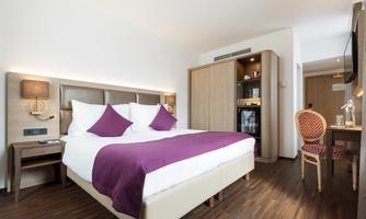 A modern hotel room with a large double bed, elegant pillows, and a comfortable armchair. The room is bright and well-equipped, with wooden floors and stylish furniture.