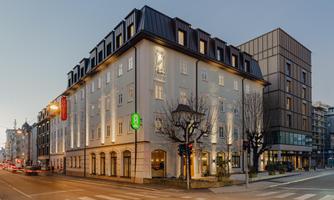 A modern building at a street corner with illuminated windows and a clear evening atmosphere. The architecture combines a classic style with modern elements.