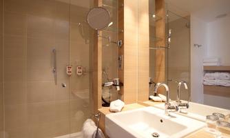 A modern bathroom with a double sink and large mirrors. The walls are tiled in a light color and towels are neatly stacked on a shelf.