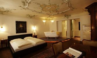 An elegant room with a double bed and an ornate ceiling pattern. In the background, there is a freestanding bathtub.