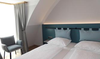 A modern bedroom with two single beds and stylish blue cushions. A cozy chair and large windows create a bright atmosphere.