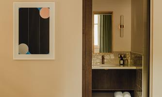 A modern bedroom with a bright color tone and a piece of artwork on the wall. In the background, access to a bathroom is visible.