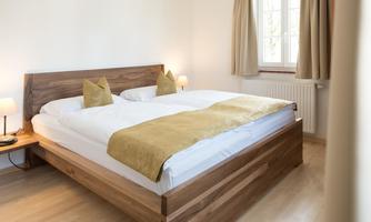 A cozy bedroom with a large wooden bed and bright curtains. Yellow cushions and a blanket are lying on the bed.