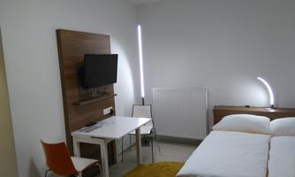 A simple room with two beds, a table, and a television on the wall. The walls are light and there is pleasant lighting.
