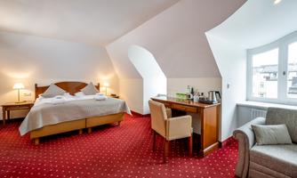 A modern hotel room with a large bed and a cozy armchair. The room is brightly and friendly designed, featuring a desk and a window.