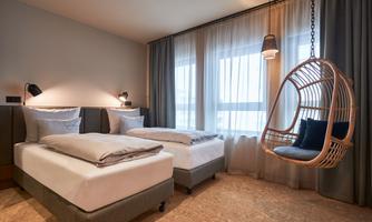 A modern bedroom with two single beds and a cozy hanging swing. Large windows with curtains provide plenty of daylight.