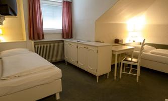 A simple room with two beds, a table, and a chair. Soft lighting and windows with curtains create a cozy atmosphere.