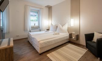 A modern bedroom with a double bed and two large pillows. The room is bright and friendly, designed with a cozy armchair and a carpet.