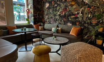 A stylish lounge with cozy seating and a decorative plant pattern on the wall. There are round tables and various cushions in warm colors.