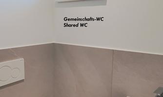 A small communal restroom with a window and a sink. The walls are light-colored and the floor is in neutral tones.