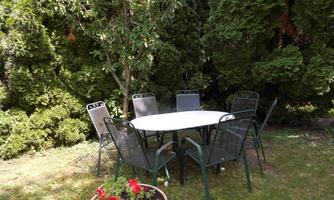 A round table with six chairs is in a green garden. In the foreground, a pot with red flowers is blooming.