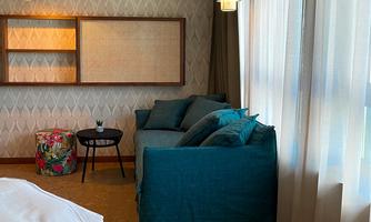 A cozy room with a large bed and a modern living area. The windows are covered with bright curtains and there is a small seating area.