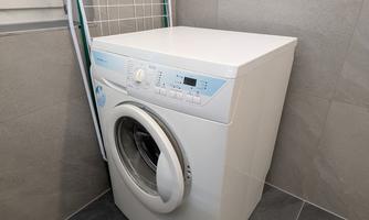 A modern washing machine stands in a room with gray tiles. In the background, a drying rack is visible.