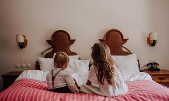 Two children are sitting on a bed and looking into the distance. The room is bright and cozy, furnished with white bedding and a striking nightstand.