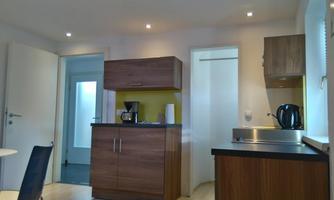 A modern kitchenette with wooden cabinets and a coffee machine. The room is brightly lit and offers a view of a door and another room.