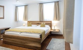 A bright bedroom with a large wooden bed and comfortable pillows. The windows are adorned with light curtains and there is a table as well as a small lamp.