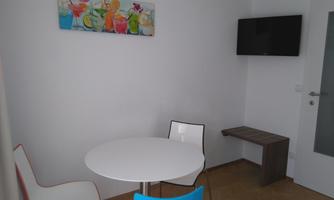 A small dining room with a round table and two chairs. A colorful painting hangs on the wall, and a television is mounted.