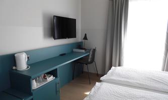 A modern hotel room with two beds, a desk, and a television. Bright colors and plenty of natural light create an inviting atmosphere.