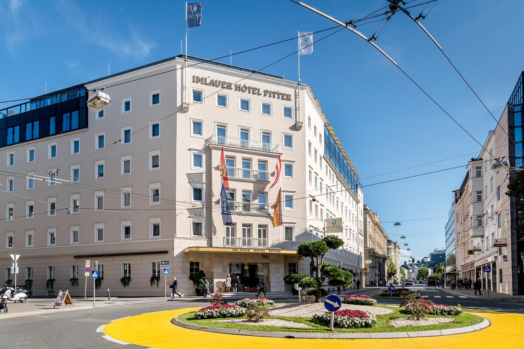 An elegant hotel building with a modern facade and colorful flower beds in the foreground. The street is well-lit and invitingly designed.