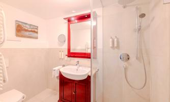 A modern bathroom with a shower and a sink. The walls are light, and there is an appealing red wooden shelf.