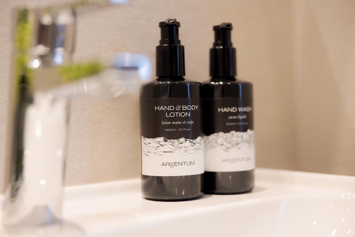Two bottles of hand and body lotion as well as hand wash are on a sink. The products are elegantly packaged and appear modern.