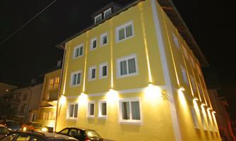 A light yellow building at night, illuminated by warm light. Several cars are parked in front of the building.
