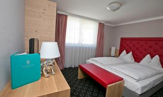 A modern bedroom with a comfortable double bed and a red bench. The room has large windows with light curtains and wooden details.