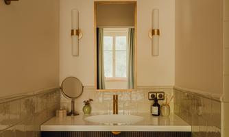 A modern bathroom with an elegant sink and golden fixtures. The large mirror reflects a window and provides plenty of natural light.