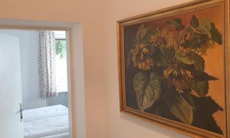 A cozy hallway with a painting of plants on the wall. In the background, a bedroom with light bedding is visible.