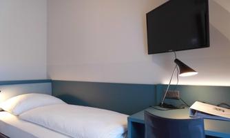 A modern single room with a cozy bed and a large TV on the wall. On the table, there is a pleasant light and some written material.