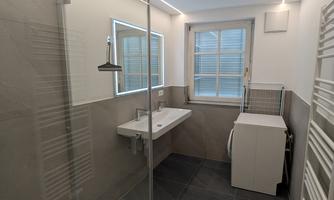 A modern bathroom with a glass shower and a simple sink. It has bright walls, large windows, and pleasant lighting.