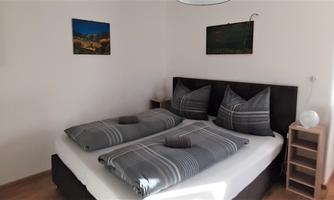 A modern bedroom with a large double bed and gray bedding. The room features pictures on the wall and a desk chair.