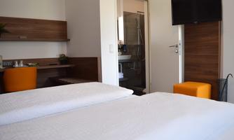 A modern hotel room with two beds and a bathroom in the background. The decor is simple with an orange stool and a television on the wall.